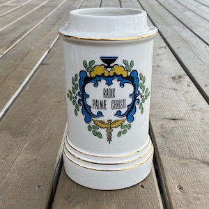 May include: White ceramic apothecary jar with gold trim and a decorative emblem featuring the text "RADIX PALMAE CHRISTI". The emblem is framed by blue and green foliage, with a yellow accent.