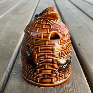 May include: A brown ceramic honey pot shaped like a beehive with a bee on the lid. The pot has a brown and gold glaze and features three bees in relief on the sides.