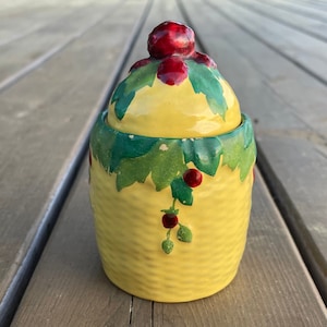 May include: A yellow ceramic jar with a lid, decorated with green leaves and red berries. The jar has a textured basket weave pattern. The lid is topped with a large red berry. A decorative kitchen item.
