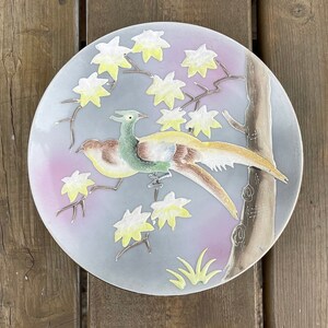 May include: A decorative ceramic plate with a bird and floral design. The plate has a light blue background with a pink gradient. The bird is green, yellow, and brown. The flowers are yellow and white.