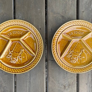 May include: Two vintage, mustard-yellow fondue plates. Each plate has three sections and a central design featuring a cow, a bottle, and the words "LA FONDUE". The plates have a rope-like border.