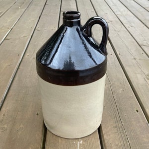 Peut inclure: Une cruche en grès avec un dessus et une anse émaillés brun foncé, et un corps de couleur beige. La cruche a un design classique et vintage, parfait pour la décoration rustique ou le stockage. La cruche mesure environ 30 cm de haut.