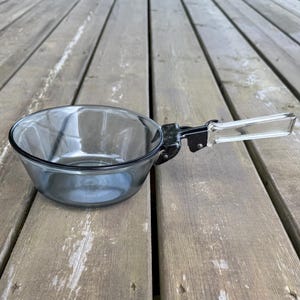 May include: A clear glass bowl with a black rim and a folding handle. The handle is clear plastic and folds over the bowl. The bowl is on a wooden surface.