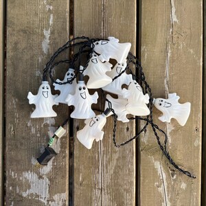 May include: A string of decorative lights featuring multiple white, ghost-shaped covers with black printed facial expressions. The ghosts are attached to a thin black electrical cord that includes a standard plug at one end for power connection.