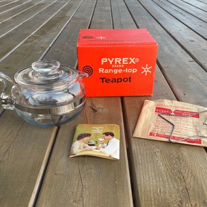 May include: A clear glass Pyrex range-top teapot with a metal band around the middle. The teapot is on a wooden surface with a red and white box and a brown paper bag with a metal heat spreader grid.