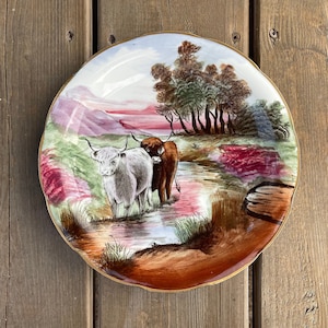 May include: A decorative plate with a painted scene of two Highland cattle in a stream. The plate has a gold rim and a landscape with trees, mountains, and a colorful sky. The cows are white and brown, typical of the breed.