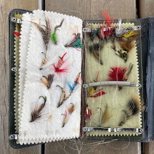 Puede incluir: Una caja de aparejos de pesca con mosca vintage abierta, que revela una selecci&oacute;n de moscas de pesca coloridas. La caja es negra con un interior verde. Las moscas est&aacute;n hechas de plumas y otros materiales, unidas a peque&ntilde;os anzuelos.