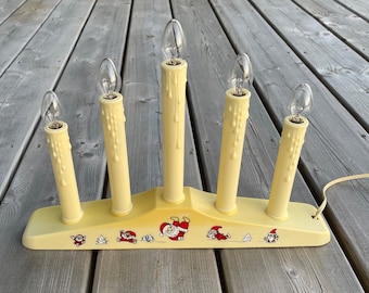 Puede incluir: Un portavelas de plástico amarillo con cinco bombillas con forma de velas. El portavelas tiene un diseño de Papá Noel y otros personajes navideños.