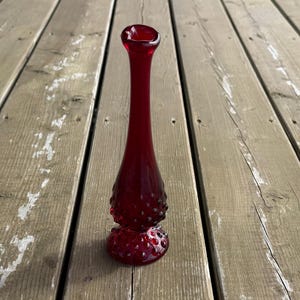 May include: A tall, ruby red glass vase with a flared top and a textured, hobnail-like pattern around the base. The vase has a slender neck and a rounded base, standing on a wooden surface.