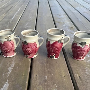 Peut inclure: Quatre mugs en céramique faits à la main, avec une base crème et un motif de rose rouge vif. Chaque mug a une anse incurvée et est posé sur une surface en bois vieilli. Les mugs mesurent environ 10 cm de haut.