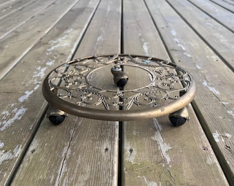 Può includere: Un supporto circolare per piante in metallo color ottone, caratterizzato da un intricato motivo a volute traforate sulla superficie superiore. Il supporto è sostenuto da tre piccole ruote piroettanti scure fissate alla base.