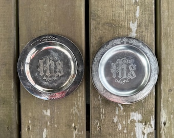 Op de afbeelding: Twee ronde zilverkleurige metalen borden met een gegraveerd IHS-monogram in het midden. De randen van de borden zijn versierd met geëtste blad- en kruismotieven. De oppervlakken vertonen tekenen van aanslag en slijtage door ouderdom.