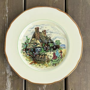 Peut inclure: Une assiette décorative avec un bord crème et un bord doré. Le centre présente une illustration détaillée d'un cottage au toit de chaume avec un jardin. L'assiette a un bord festonné et est posée sur un fond en bois.
