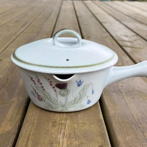 May include: A light blue ceramic teapot with a lid and a handle. The teapot features a floral design with red, blue, and green accents. The lid has a small loop handle and a vent hole. The teapot is sitting on a wooden surface.