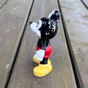 May include: A vintage Mickey Mouse figurine. The figure is black, white, red, and yellow. Mickey is wearing red shorts, white gloves, and yellow shoes. The figurine is standing on a wooden surface.