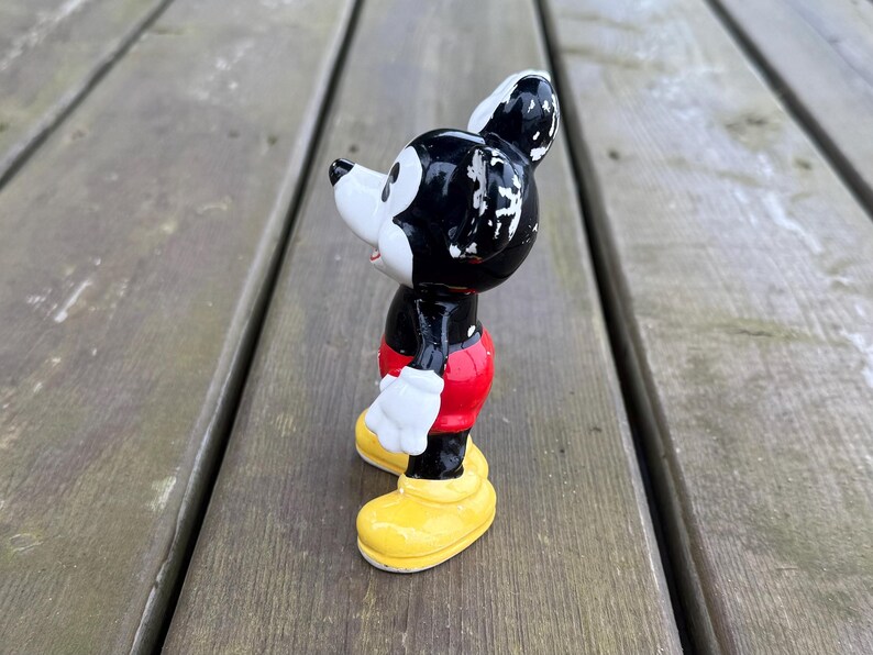 May include: A vintage Mickey Mouse figurine. The figure is black, white, red, and yellow. Mickey is wearing red shorts, white gloves, and yellow shoes. The figurine is standing on a wooden surface.