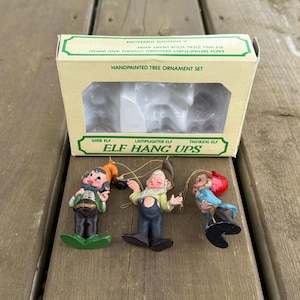 Peut inclure: Un ensemble de décorations d'arbres peintes à la main avec trois figurines d'elfes. La boîte indique "ELF HANG UPS" et comprend les phrases "Wise Elf", "Lamplighter Elf" et "Thinking Elf". Les elfes ont des tenues colorées.