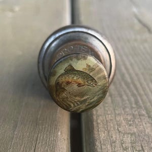 May include: A metal bottle stopper with a circular top featuring a detailed illustration of a fish. The stopper has a rustic, vintage appearance, with the word "PROTEX" visible on the side.