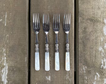 Op de afbeelding: Een set van vier zilverkleurige metalen vorken met elk vier tanden. De handvatten zijn gemaakt van een glad, iriserend wit materiaal met een decoratieve, textuurrijke metalen kraag tussen de vorkkop en de basis van het handvat.