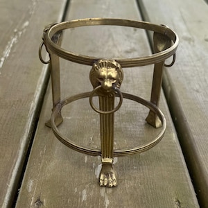 May include: A vintage brass stand with a circular top and base, supported by four vertical bars. The stand features a lion's head with a ring and a lion's paw base. The stand is designed to hold a small object.