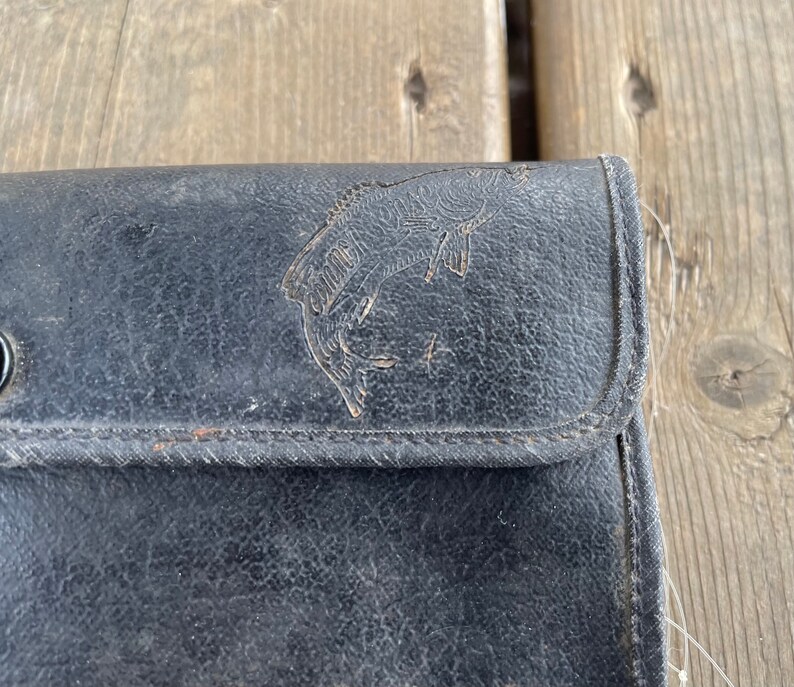Puede incluir: Estuche de aparejos de pesca de cuero negro con un dise&ntilde;o de pez en relieve. El estuche tiene un cierre de bot&oacute;n a presi&oacute;n y bordes cosidos. El cuero muestra signos de desgaste, lo que le da un aspecto vintage. Un accesorio cl&aacute;sico para pescadores.