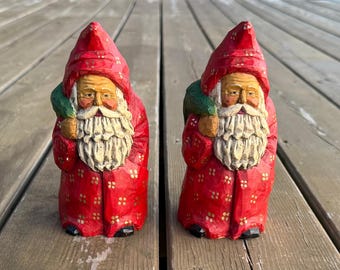 Op de afbeelding: Twee houten kerstmanbeeldjes. Elke figuur draagt een rode jas en hoed met gouden accenten. Ze hebben witte baarden en houden een groene zak over hun schouder. De beeldjes zijn gedetailleerd gesneden en staan op een houten oppervlak.