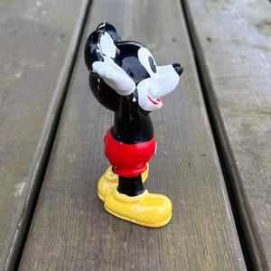 May include: A vintage Mickey Mouse figurine. The figure is black, red, white, and yellow. Mickey is wearing red shorts and yellow shoes. The figurine is standing on a wooden surface.