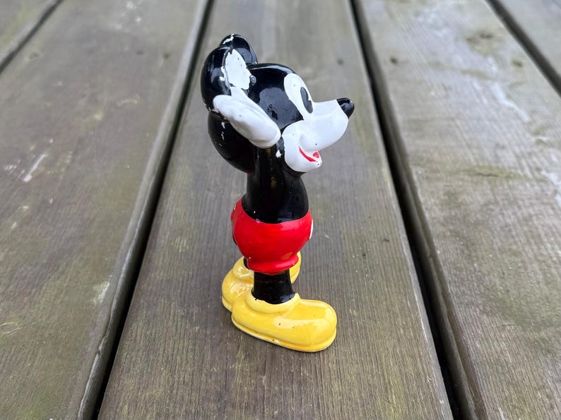 May include: A vintage Mickey Mouse figurine. The figure is black, red, white, and yellow. Mickey is wearing red shorts and yellow shoes. The figurine is standing on a wooden surface.