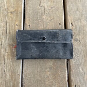 Puede incluir: Una cartera de cuero negro desgastada con cierre de bot&oacute;n a presi&oacute;n. La cartera rectangular est&aacute; cerrada, con una solapa asegurada por un solo bot&oacute;n a presi&oacute;n negro. El cuero muestra signos de desgaste. La cartera est&aacute; sobre una superficie de madera.