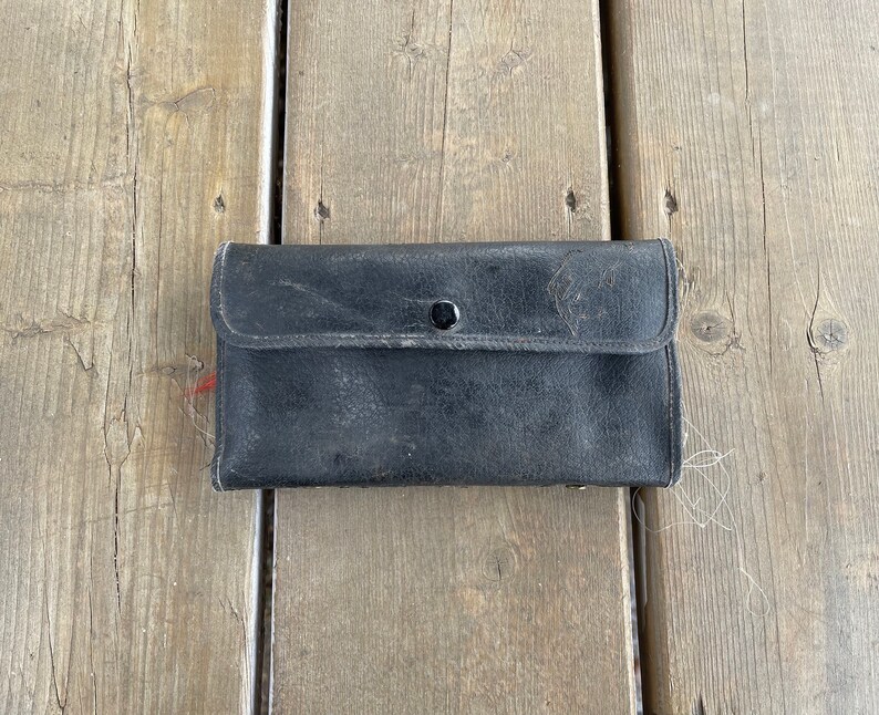 Puede incluir: Una cartera de cuero negro desgastada con cierre de bot&oacute;n a presi&oacute;n. La cartera rectangular est&aacute; cerrada, con una solapa asegurada por un solo bot&oacute;n a presi&oacute;n negro. El cuero muestra signos de desgaste. La cartera est&aacute; sobre una superficie de madera.