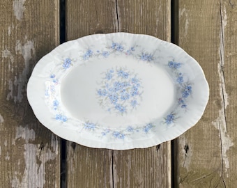 Può includere: Un piatto da portata ovale in porcellana bianca con bordo smerlato e un delicato motivo floreale di piccoli fiori blu con foglie verdi, disposti in un grappolo centrale e in un bordo continuo lungo tutto il perimetro.