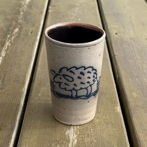 May include: A tall, cylindrical ceramic cup with a sheep illustration. The cup is a light beige colour with a dark blue sheep design. The interior of the cup is dark brown.