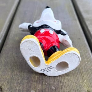 May include: A ceramic Mickey Mouse figurine in a lying down pose. The figure has a white head, black ears, and a red torso with white buttons. It wears black gloves, and yellow shoes. The bottom of the shoe reads "WALT DISNEY PRODUCTIONS, JAPAN."