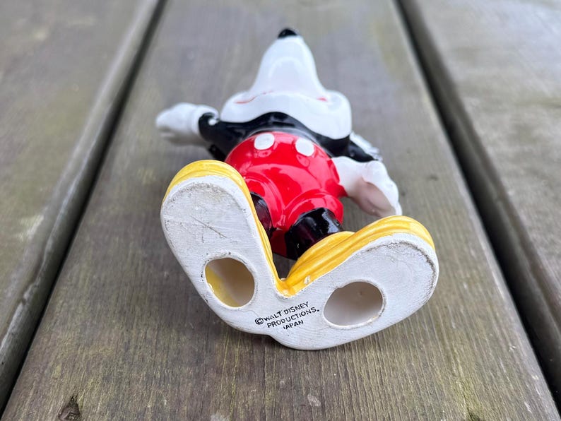 May include: A ceramic Mickey Mouse figurine in a lying down pose. The figure has a white head, black ears, and a red torso with white buttons. It wears black gloves, and yellow shoes. The bottom of the shoe reads "WALT DISNEY PRODUCTIONS, JAPAN."