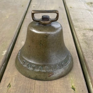May include: A bronze bell with a handle and the word "CANADIAN" embossed around the base. The bell is set on a weathered wooden surface, showcasing its aged appearance and vintage charm.