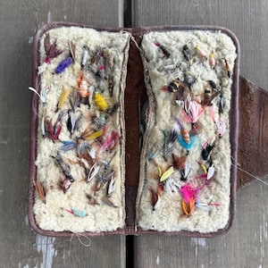 May include: Open, vintage leather fly fishing case filled with colorful artificial flies. The case is open, revealing a cream-colored, textured interior. The flies are in various colors, including red, yellow, blue, and pink. The case is on a weathered wooden surface.