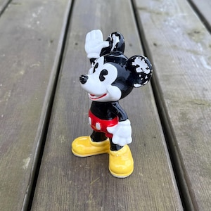 May include: A vintage Mickey Mouse figurine waving. The figure is black, with white gloves and yellow shoes. Mickey's shorts are red with white buttons. The figurine stands on a wooden surface. The figure is approximately 10 cm tall.