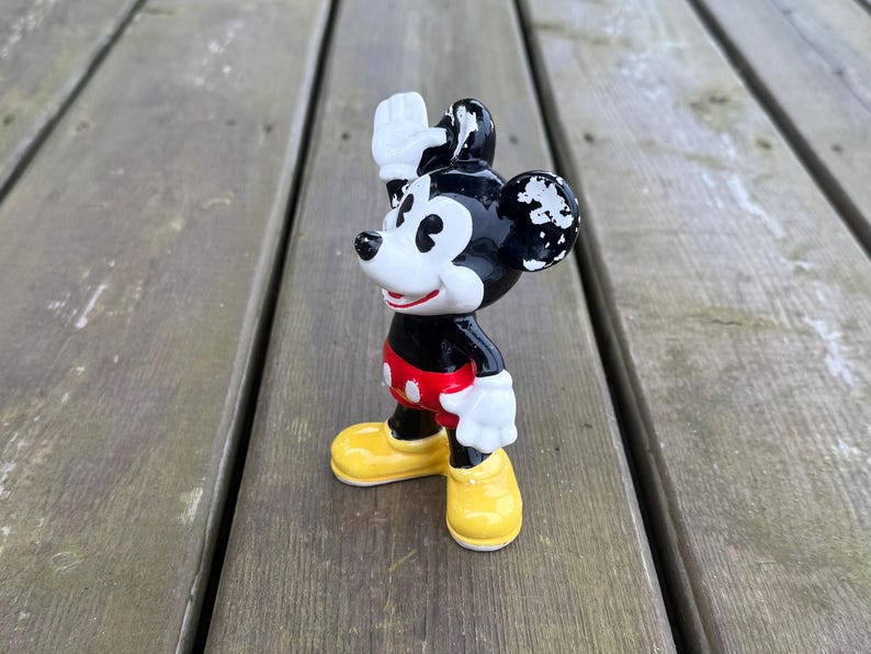 May include: A vintage Mickey Mouse figurine waving. The figure is black, with white gloves and yellow shoes. Mickey's shorts are red with white buttons. The figurine stands on a wooden surface. The figure is approximately 10 cm tall.