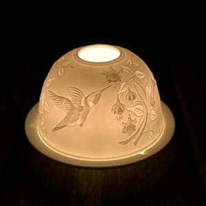 May include: A white porcelain lamp shade with a hummingbird design. The shade is dome-shaped with a circular base and features an embossed hummingbird and floral pattern. The lamp is lit from within, creating a soft glow.
