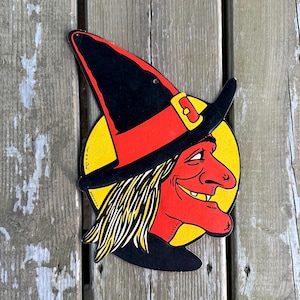 May include: A flat, die-cut wall decoration featuring a profile illustration of a witch with red skin, a long nose, and a pointed black hat with a red band and yellow buckle. The witch has stringy yellow hair and is set against a bright yellow circle.