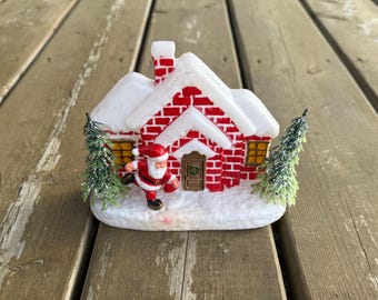 Op de afbeelding: Een miniatuur vakantiehuisje met rode bakstenen muren, een wit besneeuwd dak en een schoorsteen. Een klein kerstmanfiguurtje staat voor de bruine deur, geflankeerd door twee groene dennenbomen op een witte besneeuwde ondergrond.