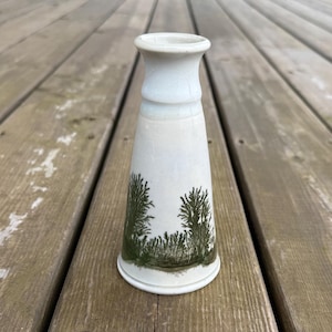 May include: A white ceramic vase with a flared base and a slightly wider top. The vase features a hand-painted design of green trees and grass. The vase is approximately 18 cm tall.