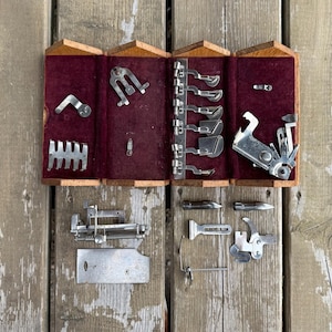Peut inclure: Une collection d'accessoires et de pieds-de-biche pour machine à coudre en métal argenté. Plusieurs composants sont organisés dans un coffret en bois doublé de velours bordeaux, tandis que d'autres pièces métalliques sont disposées sous le coffret.