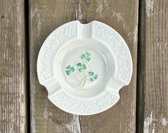 Può includere: Un posacenere rotondo in ceramica color bianco sporco con un disegno centrale di trifoglio dipinto in verde. Il bordo è decorato con un motivo a intreccio celtico in rilievo e include tre tacche per appoggiare oggetti sul bordo.