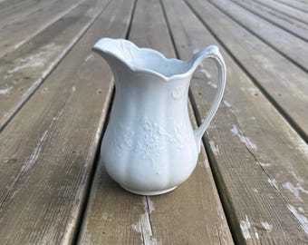 Può includere: Una brocca in ceramica bianca con bordo smerlato, manico curvo e un sottile motivo floreale in rilievo sul corpo. La brocca presenta una base arrotondata e un beccuccio svasato, con una finitura lucida e dettagli verticali scanalati.