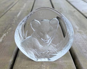 May include: A clear glass sculpture of a lion cub. The cub is resting inside a rounded, clear glass frame. The sculpture is set on a wooden surface.
