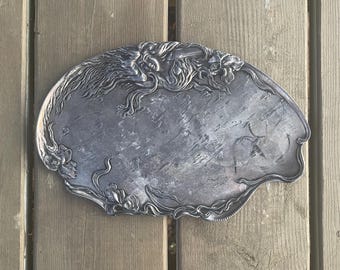 Könnte beinhalten: Ein ovales Serviertablett aus Silber mit einem verzierten Design aus Blumen und Blättern. Das Tablett hat eine strukturierte Oberfläche und einen leicht erhöhten Rand.