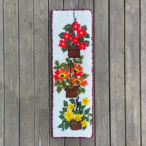 Peut inclure: Une tenture murale rectangulaire et texturée représentant trois paniers de fleurs suspendus. Le panier supérieur contient des fleurs rouges, le milieu des fleurs orange et jaunes, et le bas des fleurs jaunes. Le fond est blanc avec une bordure rouge.
