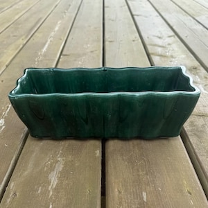 Peut inclure: Un pot de fleurs rectangulaire en céramique vert foncé avec un bord ondulé. Le pot est vide et repose sur une surface en bois vieilli. Le pot mesure environ 30 cm de long.