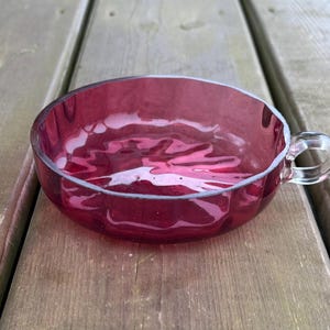 May include: A ruby-red glass teacup with a handle. The cup has a round bowl shape and sits on a wooden surface. The glass is transparent, allowing light to pass through and reflect off the surface.